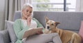 Senior woman writing notes in journal, playing crossword puzzle, sitting on sofa with pet dog. Elderly lady relaxing in