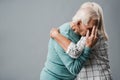 senior woman with grey hair hugging upset husband covering face on grey.