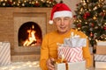 Senior wrinkled man sitting in living room with Christmas decoration and holding stack of present boxes, looking ar camera, posing