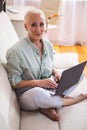 Seniors and Technologies Concepts. Smiling Relaxing Caucasian Senior Woman Posing With Laptop And Listening To The Music on Couch