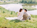 Sensitive outdoor back view of the hugging newlyweds sitting on the meadow and looking at each other.