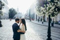 Sensitive wedding portrait in the sunny street. Handsome groom is softly touching the hair of his brunette bride near