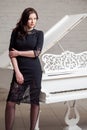 Sensual brunette woman in lace black classic dress standing near white piano and looking at camera Royalty Free Stock Photo