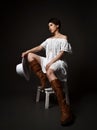 Romantic cowgirl, brunette woman in rustic clothing white lace summer dress and brown high boots sits on chair tired