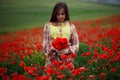 Handsome girl wearing in summer floral dress, seated in poppies field, holdings a bouquet of flowers, looks down