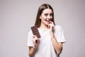 Beautiful young woman biting a bar of milky chocolate playfull look on camera before eat isolated on gray. Health concept.