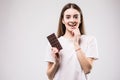 Beautiful young woman biting a bar of milky chocolate playfull look on camera before eat isolated on gray. Health concept.