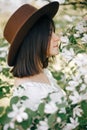 Sensual portrait of beautiful hipster woman in hat smelling white flowers in spring. Stylish calm boho girl posing in blooming