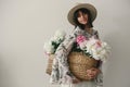 Sensual portrait of boho girl holding pink and white peonies in rustic basket. Stylish hipster woman in hat and bohemian floral