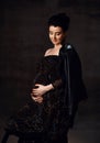 Sensual pregnant noble woman in black off-shoulder dress and leather jacket stands holding hand at her belly, feeling love Royalty Free Stock Photo
