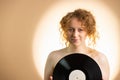 Sensual redhead young woman covering her naked breast with vynil record round form black color posing over bright yellow Royalty Free Stock Photo