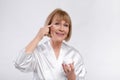 Sensual senior lady in silk bath robe applying face cream and smiling at camera over light studio background Royalty Free Stock Photo