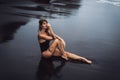 Sensual sexy girl in swimsuit with wet hair sits on black sand beach Royalty Free Stock Photo