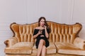 Sensual woman in trendy black dress drinking wine while sitting on sofa. Indoor portrait of worried curly girl posing Royalty Free Stock Photo