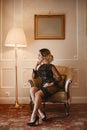 Sensual young businesswoman with trendy hairstyle in a stylish dress sits in armchair at vintage interior. Model woman