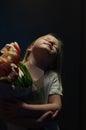 Sensuous portrait of a cute blonde young girl with tulips on a dark background under a ray of light.