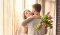 Sentimental woman with flowers embracing her loving boyfriend near window at home, panorama. Space for text