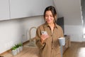 Serene asian woman stands in the kitchen with cup of coffee, checking messages