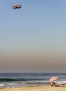 Serene California beach scene, with lone beach goer under umbrella and vintage airplane flying above.