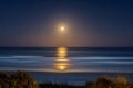 Serene ocean view at night with full moon casting glowing reflection over calm waves and sandy beach surrounded by coastal Royalty Free Stock Photo