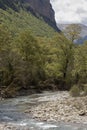 a serene river flowing through a rocky bed, surrounded by lush green trees and towering mountains under cloudy skies Royalty Free Stock Photo