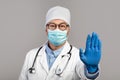 Serious adult asian doctor in white coat, glasses and protective gloves shows stop sign with hand