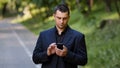 Serious adult caucasian man user stand outdoors in park wears black formal suit hold mobile telephone scrolling slide Royalty Free Stock Photo