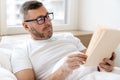 Serious adult unshaven man reading book while lying in bed