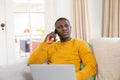 Serious african american man using laptop and talking on smartphone sitting on couch at home Royalty Free Stock Photo