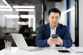 Serious Asian businessman sitting in the office at the desk with a laptop and using a mobile phone, texting, typing Royalty Free Stock Photo