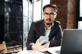 Serious asian businessman sitting at table with laptop and looking at phone while holding it in hand Royalty Free Stock Photo