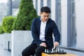 Serious asian man playing chess outside, businessman thinking playing chess sitting on bench near office center Royalty Free Stock Photo