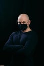 A serious bald man in a black medical mask crossed his arms over his chest. Portrait of a man on a dark background. Royalty Free Stock Photo