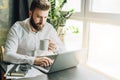 Serious bearded businessman is sitting at table, working on computer, drinking coffee. Man checking email, planning. Royalty Free Stock Photo