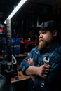 Serious bearded cycling repairman in cap holding wrench standing with crossed arms by bicycle in repair workshop with