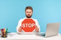 Serious bearded man office worker holding red stop sign board sitting at workplace with laptop, rejecting sexual harassment at Royalty Free Stock Photo