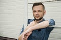 Serious bearded young man in denim shirt is standing by the wall behind ladder and looking straight into the camera Royalty Free Stock Photo