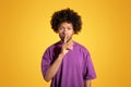 Serious black adult curly man in purple t-shirt presses finger to lips, making shh sign