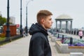 A serious blond man in a leather jacket stands on the city embankment against the backdrop of a rotunda. Side view. Blur the