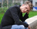 Serious blond young man in jeans and jacket, sitting outdoors