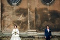Serious bride and groom stand under round steel windows of an old cathedral