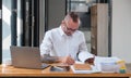 Serious business man using laptop sitting at the table in a home office, looking at the paper, communicating online Royalty Free Stock Photo