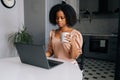 Serious busy African American business woman typing on laptop looking to device screen and drinking coffee or tea in Royalty Free Stock Photo