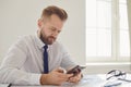 Serious busy businessman reads message on the phone at a table with a computer in the office. Royalty Free Stock Photo