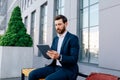 Serious busy confident young caucasian male with beard in suit typing on tablet, surfing