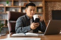 Serious busy mature chinese businessman with computer reads message on smartphone and drinks coffee Royalty Free Stock Photo