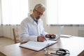 Serious busy mature doctor filling documents, sitting at work desk