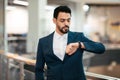 Serious calm attractive young arab ceo man with beard in suit looks at smart watch wait for client in office interior