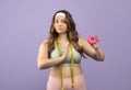Serious caucasian plus size young lady in sportswear refuses donut, gesturing on purple background