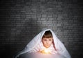 Serious child with flashlight hiding under blanket